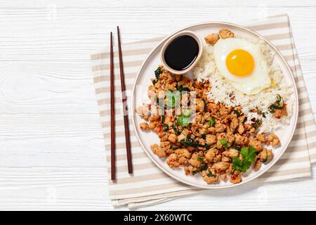 Pad Krapow Gai, Pad Gaprao, Pad kra Pao, Basilikum-Hühnchen-Rührbraten mit Reis und Spiegelei auf Teller mit Essstäbchen auf weißem Holztisch, thai-Küche, Stockfoto