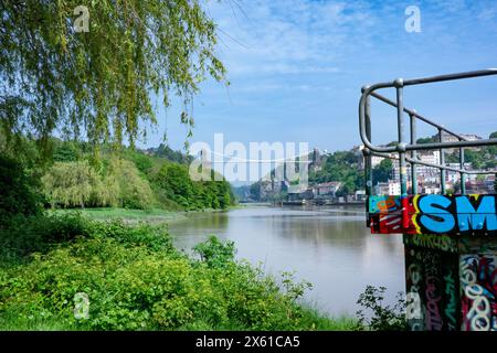 Ein Blick bei Flut entlang des Flusses Avon, Bristol, Großbritannien in Richtung Clifton Suspension Bridge. Eine mit Graffiti bedeckte Wand und eine Grasbank im Vordergrund Stockfoto