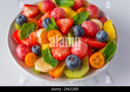 Sommer-Obstsalat in einer Schüssel auf weißem Marmorhintergrund. Gesunde Ernährung Essen zum Frühstück. Gemischte Früchte, Beeren und Minze für Diätessen Stockfoto