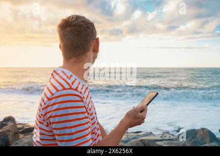 Rückansicht des kaukasischen jungen Mannes, der mit Kopfhörern musc über das Mobiltelefon an der Küste hört und den Sonnenuntergang genießt. Hübscher Mann draußen Stockfoto