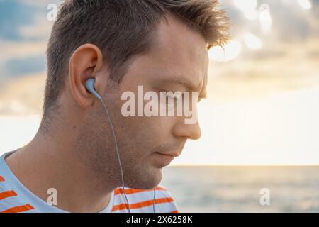 Porträt eines kaukasischen jungen Mannes, der an der Küste Kopfhörer trägt und Musik hört. Hübscher Mann draußen mit Kopfhörern am Strand Stockfoto