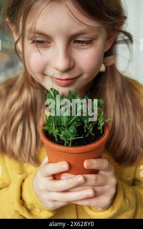 Ein Kind in einem leuchtend gelben Pullover hält zart einen Terrakotta-Topf mit einer grünen Pflanze. Sie sieht die Pflanze mit einem sanften Lächeln und einem Stockfoto