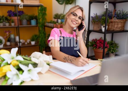 Junge blonde Frau Floristin im Gespräch auf Smartphone Schreiben auf Notebook im Blumenladen Stockfoto