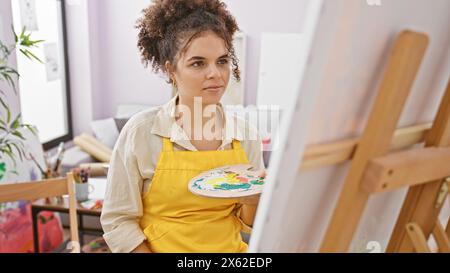 Eine junge hispanische Frau mit lockigen Haaren konzentriert sich auf die Malerei in Innenräumen und strahlt Kreativität und Konzentration in einem Kunststudio aus. Stockfoto