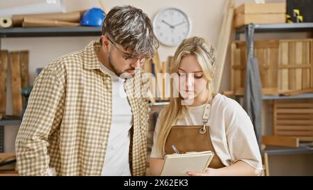 Ein Mann und eine Frau, wahrscheinliche Partner, arbeiten in einer Holzwerkstatt zusammen und konzentrieren sich auf die Projektplanung und Notizen. Stockfoto