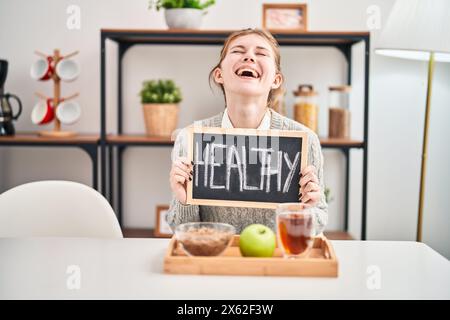 Bauchlächende junge blonde Schönheit, die zu Hause ein gesundes Frühstück genießt - ihr ansteckender, himmelhoher Geist trifft an die Decke, während sie ihre Tafel greift, Stockfoto