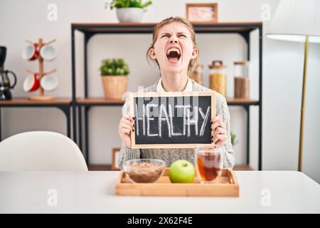 Wütende junge blonde Frau, die frustriert beim gesunden Frühstück zu Hause schreiend und wütend die Tafel hält Stockfoto