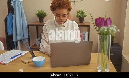 Fokussierte afroamerikanische Frau, die an einem Laptop in einem gemütlichen Heimbüro mit Blumen arbeitet. Stockfoto