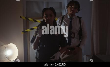 Eine Frau und ein Mann, beide Polizisten, untersuchen einen dunklen Tatort drinnen mit dramatischer Beleuchtung Stockfoto