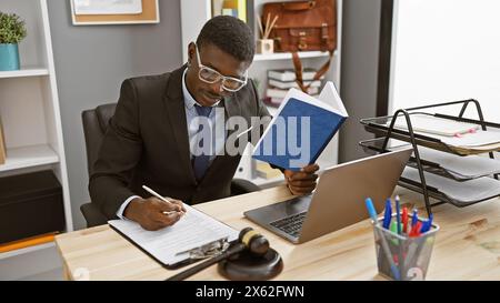 afroamerikanischer Geschäftsmann im Büro, der Dokumente analysiert und Notizen macht, mit Laptop und Hamel sichtbar. Stockfoto