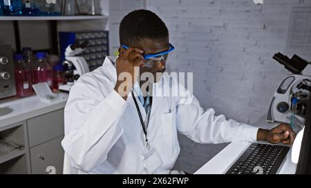 Ein fokussierter afrikanischer Mann mit Schutzbrille arbeitet an einem Computer in einem weißen Labor, was auf eine wissenschaftliche Forschungsumgebung hindeutet. Stockfoto