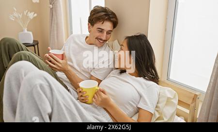 Ein fröhliches, interrassisches Paar genießt gemeinsam Kaffee in einem gemütlichen Schlafzimmer, das Wärme und Intimität weckt. Stockfoto