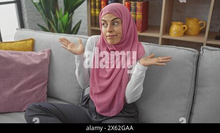 Eine junge Frau in einem rosa Hijab, die Unsicherheit ausdrückt, während sie auf einem Sofa in einem modernen Wohnzimmer sitzt. Stockfoto