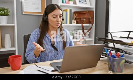 Hispanische Frau, die ein Headset trägt, im Büro mit Laptop- und Schreibtischzubehör diskutiert, was Professionalität und Kommunikation hervorruft. Stockfoto