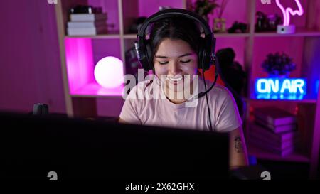 Hispanische Frau, die nachts in einem farbenfrohen Raum spielt, beleuchtet von Neonlichtern und trägt Kopfhörer. Stockfoto