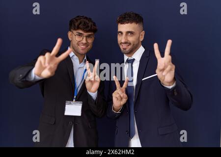 Zwei hispanische Geschäftsmänner in Geschäftskleidung lächelnd in die Kamera blicken und zeigen Finger beim Siegeszeichen. Nummer zwei. Stockfoto