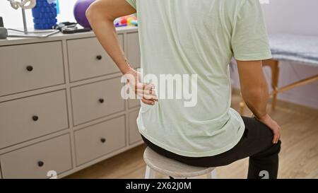 Ein junger Mann in einer Reha-Klinik hält sich zurück und deutet auf Schmerzen in einer klinischen Inneneinrichtung hin. Stockfoto