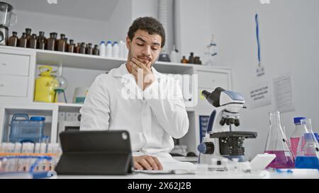 Ein besinnlicher Mann in einem Labormantel analysiert Proben in einem modernen Labor, umgeben von wissenschaftlichen Geräten. Stockfoto