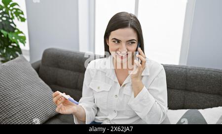 Junge hispanische Frau lächelt, während sie einen Schwangerschaftstest hält und am Telefon in einem modernen Wohnzimmer spricht. Stockfoto
