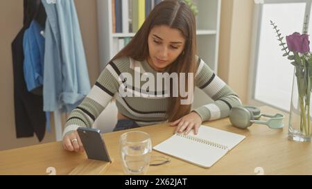 Eine junge hispanische Frau liest aufmerksam von einem Smartphone an einem Holztisch in einem gut beleuchteten Wohnraum, mit einem Notebook und einem Glas Wasser in der Nähe. Stockfoto