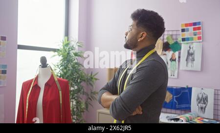 Ein kontemplativer schwarzer Mann mit überkreuzten Armen, der ein Maßband in einer Schneiderei trägt, umgeben von Designskizzen und Stoffmustern. Stockfoto