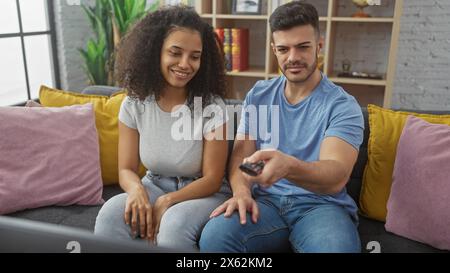 Ein junger Mann und eine junge Frau sitzen dicht auf einem gemütlichen Sofa, genießen ihre gemeinsame Zeit in einem modernen Wohnzimmer und schauen Fernsehen. Stockfoto