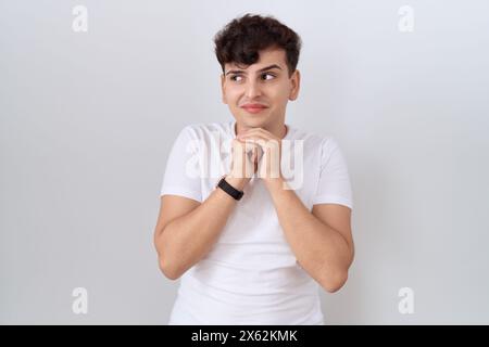 Junger, nicht binärer Mann mit lässigem weißem T-Shirt lachend nervös und aufgeregt mit Händen am Kinn zur Seite schauend Stockfoto
