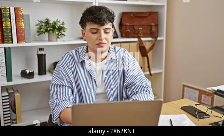 Ein junger Mann arbeitet nachdenklich an einem Laptop, umgeben von Büroregalen mit Büchern und Dekor. Stockfoto