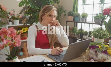 Eine nachdenkliche junge Frau in einem Blumenladen, umgeben von bunten Blumen, arbeitet drinnen an ihrem Laptop. Stockfoto
