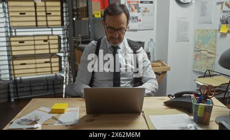 Reifer bärtiger Mann, der im Detektivbüro mit Karte und Dokumenten am Laptop arbeitet. Stockfoto