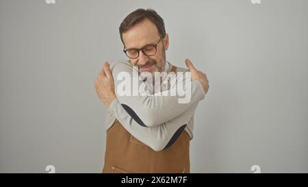 Ein bärtiger Mann mittleren Alters in lässiger Kleidung umarmt sich vor einem isolierten weißen Hintergrund und drückt Komfort oder Selbstliebe aus. Stockfoto