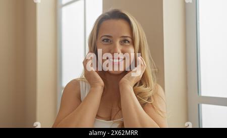 Eine lächelnde blonde Frau berührt ihr Gesicht sanft, während sie drinnen sitzt, und strahlt lässige Eleganz und Schönheit aus. Stockfoto