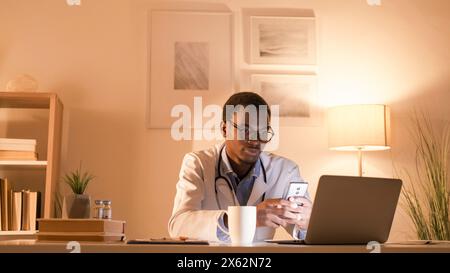Arzt online. Mobile Telemedizin. Internetberatung. Intelligenter männlicher Facharzt, der mit Telefon-Laptop für Online-Chat im modernen Arbeitsplatz-Innenraum mit Kopierraum arbeitet. Stockfoto