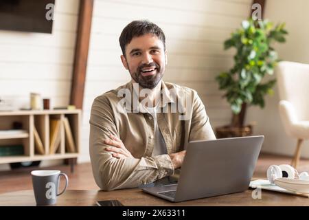 Fröhlicher Mann sitzt am Holztisch im gut beleuchteten Home Office Stockfoto