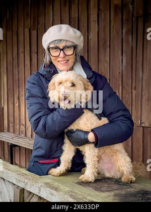 Porträt einer Reifen kaukasischen Frau in Wintermantel und Wollmütze, die einen blonden Kakapohund umschließt. Stockfoto