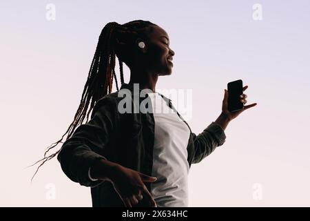 Eine junge Frau mit Zöpfen tanzt selbstbewusst in einem Studio mit Ohrstöpseln und Smartphone in der Hand. Sie feiert die Liebe von Gen Z zu Musik und Technologie, s Stockfoto