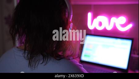 Junge Freelancerin arbeitet von zu Hause aus auf Laptop, Kopfhörer an, neonrosa Lampe darüber. Blick von hinten, zeigt moderne Fernarbeiten Stockfoto