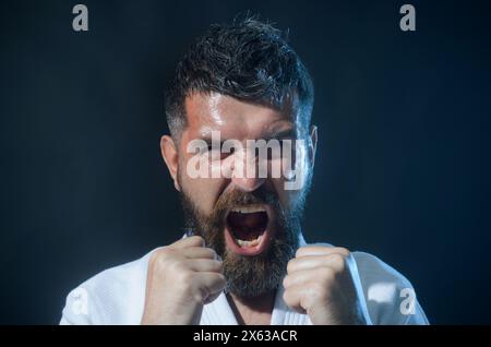 Wütender schreiender bärtiger Mann im weißen Kimono in Kampfposition. Karate-Kämpfer, der sich auf den Wettbewerb vorbereitet. Muskuläres männliches Fitness-Modell bereit für Stockfoto