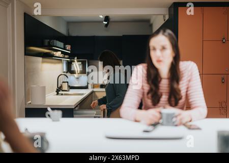 Konzentrieren Sie sich auf eine moderne Kücheneinrichtung, in der eine Frau an der Theke arbeitet und eine andere unfokussiert im Vordergrund sitzt, was das tägliche Leben widerspiegelt. Stockfoto