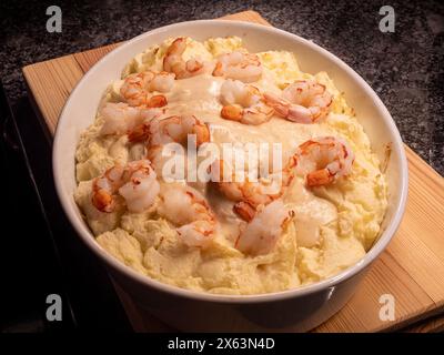 Ein dampfender weißer Auflauf gefüllt mit prallen Garnelen, gebadet in einer leichten Weißweinsauce, eingebettet in cremige Kartoffelpüree, alles auf einem Holzbrett. Stockfoto