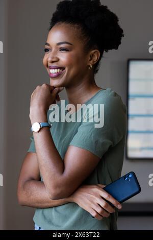 Im Büro eine afroamerikanische Geschäftsfrau, die ihr Smartphone hält, wegblickt und lächelt Stockfoto