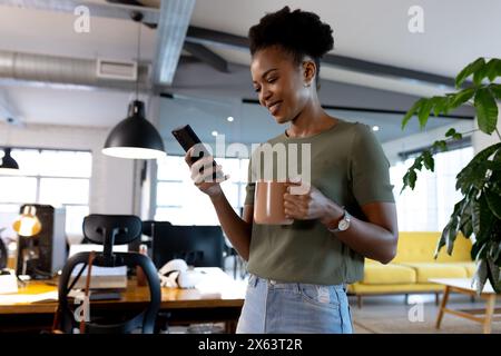 Im Büro, eine afroamerikanische Geschäftsfrau, die Kaffee hält und auf das Telefon schaut Stockfoto