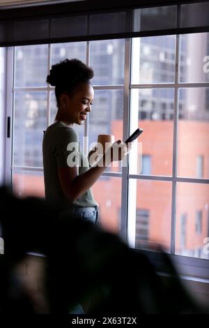 Im Büro eine afroamerikanische Geschäftsfrau, die ihr Smartphone am Fenster hält Stockfoto