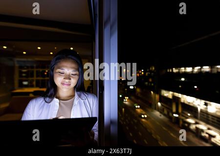 Im Büro, eine Geschäftsfrau mit birassischer Herkunft, die ein Tablet am Fenster untersucht, bis spät arbeitet, einen Kopierraum hat Stockfoto
