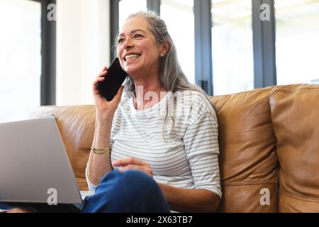 Zu Hause, kaukasische reife Frau, die mit dem Smartphone spricht, mit dem Laptop sitzt Stockfoto