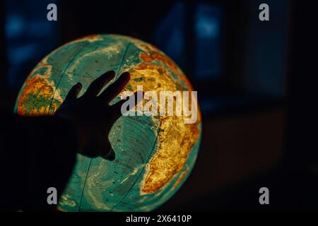 Die Welt erkunden: Eine Hand reicht zu einer glühenden Kugel in einem dunklen Raum. Bildungsreise: Leuchtkugel und Suche nach Ländernamen Stockfoto