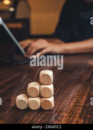 Pfeil, der auf Holzstäben springt, Balkendiagramm-Stufen auf dem Schreibtisch, während Geschäftsmann mit Laptop arbeitet, vertikaler Stil. Wachstum des Geschäftsmarktes, Aufwand, Stockfoto