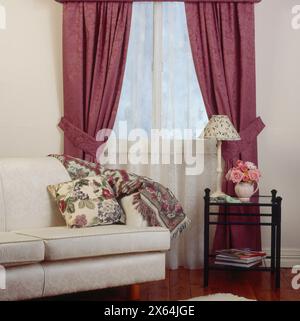 INNENANSICHT DES LOUNGE-ZIMMERS MIT FENSTER MIT VORHÄNGEN, LOUNGE, KISSEN, ÜBERWURF UND KLEINEM BEISTELLTISCH. Stockfoto