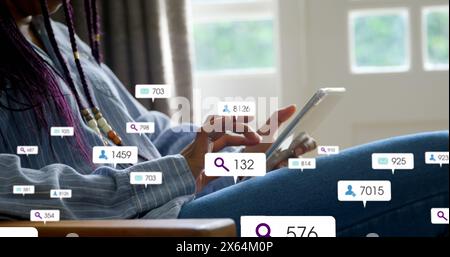 Bild von Social-Media-Ikonen, die über dem mittleren Teil einer Frau schweben, die zu Hause ein digitales Tablet verwendet Stockfoto