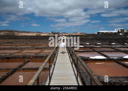 Eine Frau mit Strohhut geht über eine Brücke über ein Salzfeld. Der Himmel ist blau und die Sonne scheint Stockfoto